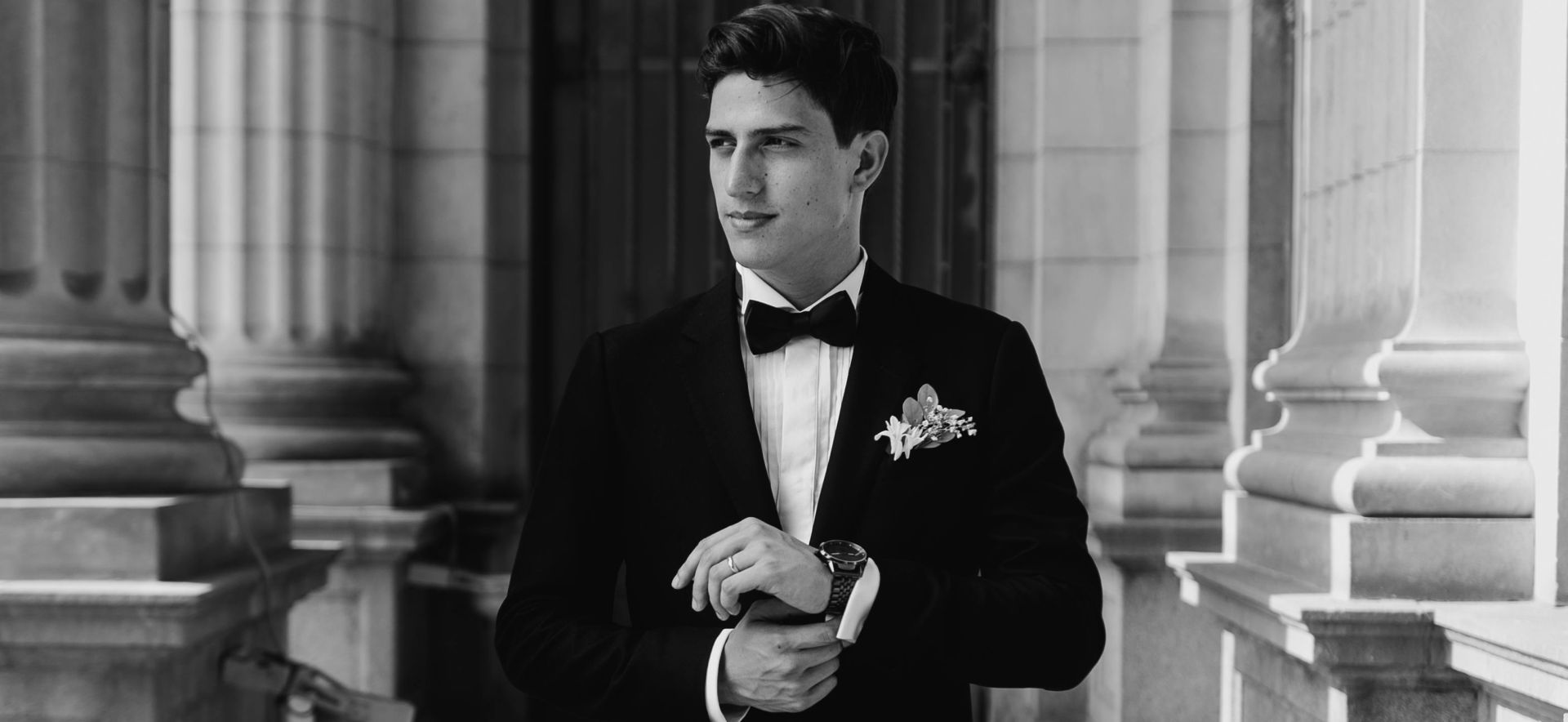 A sophisticated bridegroom adjusts his watch, standing amidst classical columns in elegant black and white.